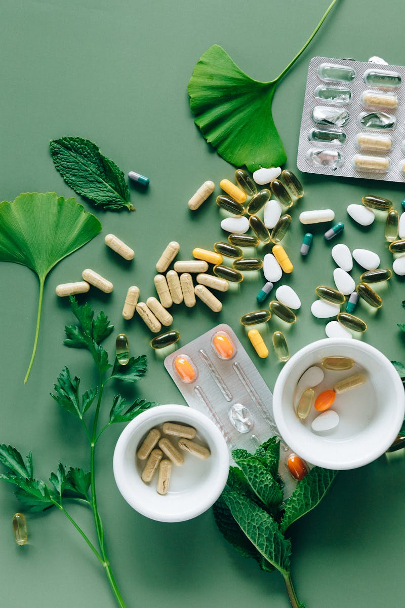 Flat lay of assorted pills and herbs on green surface, symbolizing health and wellness.